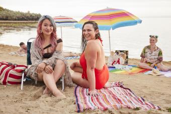 Rora and Akii are sitting on the beach. Akii is facing the camera, and is wearing a beach shawl. She has pink hair and is smiling at the camera. Rora is in a red bathing suit and is turning towards the camera. Grace is in the background smiling. There is a rainbow beach umbrella and beach towels near them.