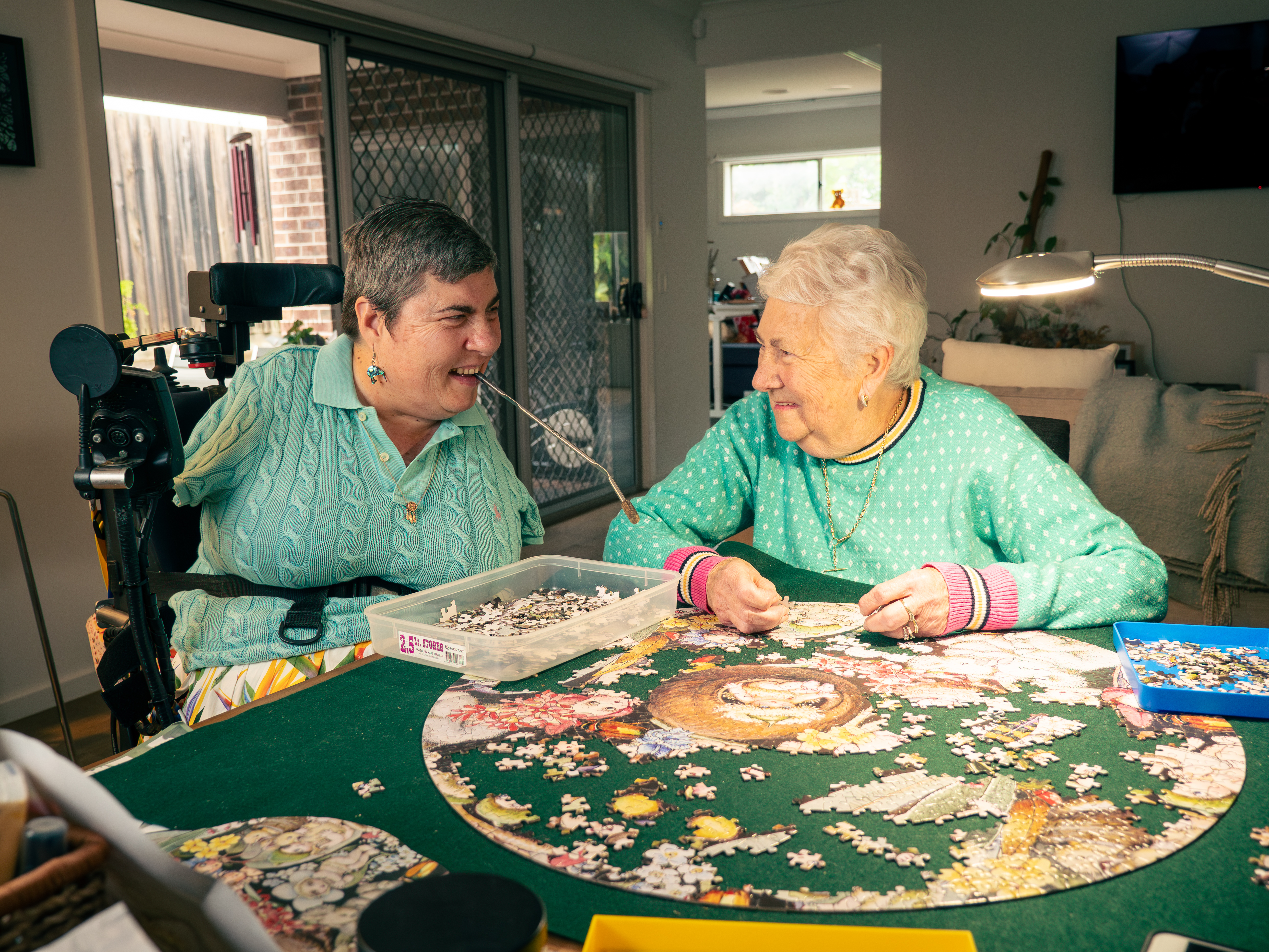 A picture of two women sitting at a table smiling while they are completing.a jigsaw puzzle.They are both wearing green tops and the woman on the left is sitting in a wheelchair and has no arms. 