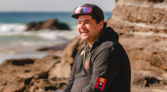 Jesse is sitting on the rocks at the beach with the waves rolling in the background. He is wearing a baseball hat and a black hooded jumper. He is smiling away from camera.