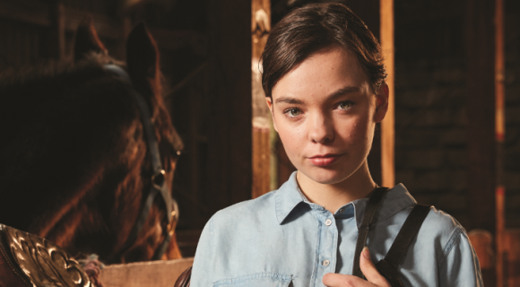 Chloe is wearing a blue button up shirt and is holding a horse saddle. She is standing in the stables next to a horse and is smiling at the camera.